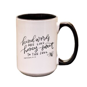 May include: White ceramic mug with a black handle and black interior. The mug features a black and white illustration of a bee and the text "kind words are like honey-sweet to the soul Proverbs 16:24".