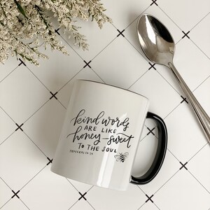 May include: White ceramic mug with a black handle. The mug has a black design that says "Kind words are like honey-sweet to the soul" with a bee graphic and the text "Proverbs 16:24".