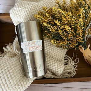 May include: A stainless steel tumbler with a white sticker that reads "nevertheless she persisted". The tumbler has a black Yeti logo on the bottom.