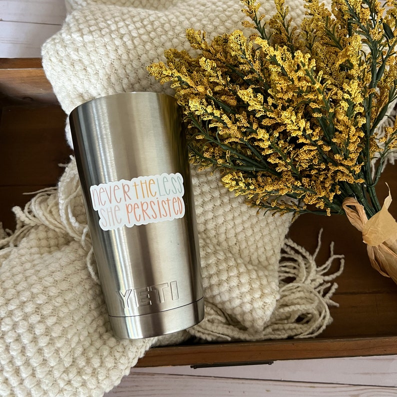 May include: A stainless steel tumbler with a white sticker that reads "nevertheless she persisted". The tumbler has a black Yeti logo on the bottom.