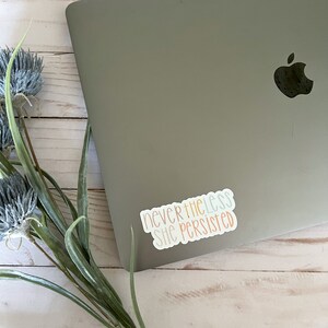 May include: A sticker with the text "nevertheless she persisted" in a rainbow colour scheme, placed on a silver laptop.