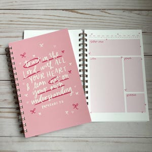 May include: Pink spiral bound journal with a quote from Proverbs 3:6, "Trust in the Lord with all your heart and lean not on your own understanding." The journal has a daily planner section with sections for today's verse, notes, prayer, and gratitude.