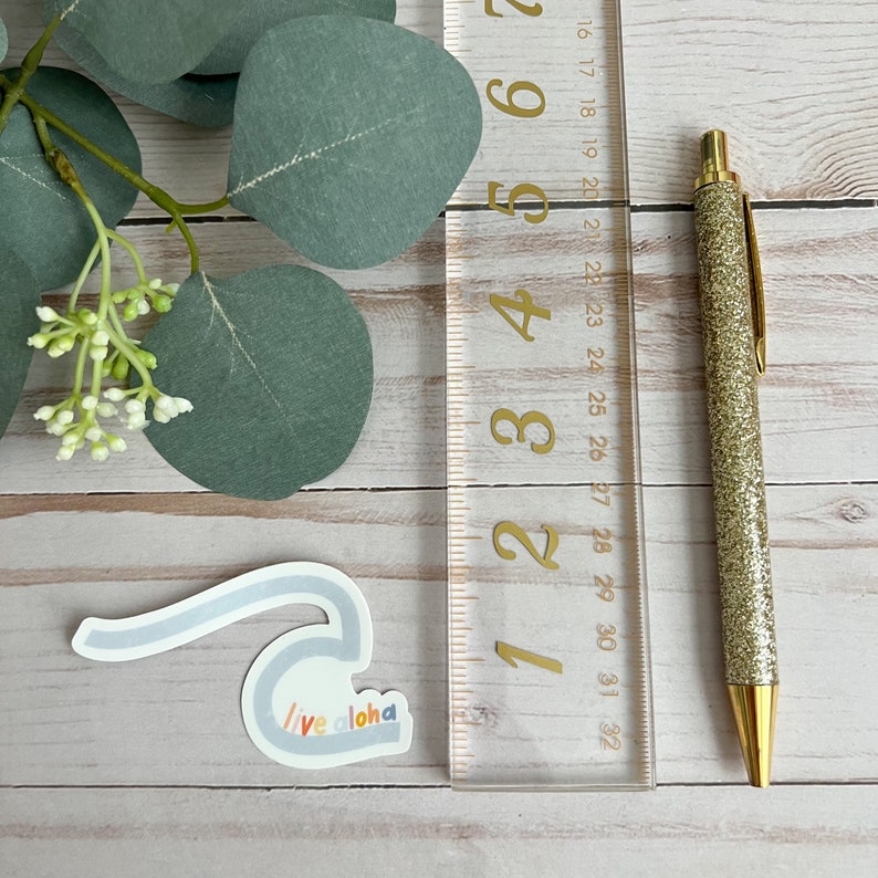 May include: A light blue sticker with a wave design and the text "Live aloha" in rainbow colors. The sticker is placed on a wooden surface next to a ruler and a gold pen.