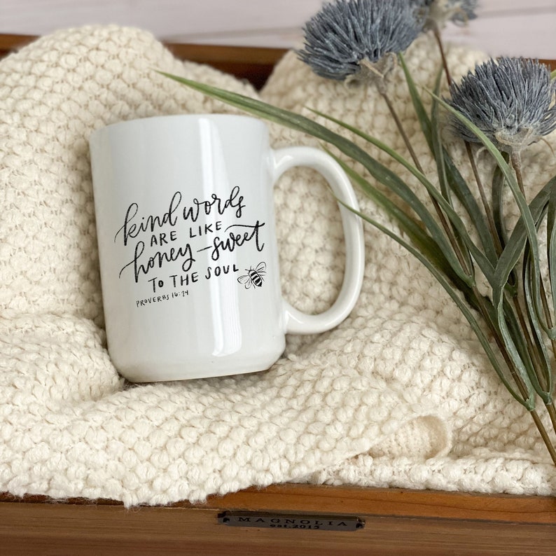 May include: White ceramic mug with a black handwritten quote that reads "Kind words are like honey-sweet to the soul" with a small bee illustration and the scripture reference "Proverbs 16:24".