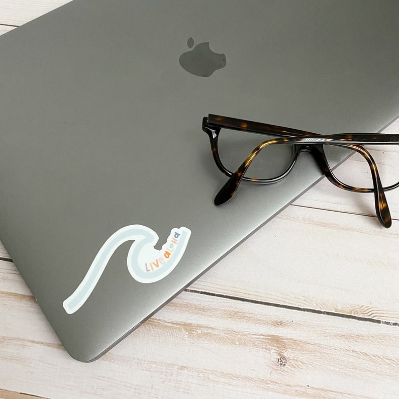 May include: A silver laptop with a sticker that says "Live Aloha" in rainbow colors on a wave design. A pair of tortoiseshell glasses are on top of the laptop.