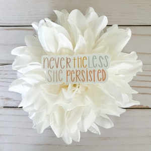 May include: A white flower with a sticker that reads "nevertheless she persisted" in colourful lettering.