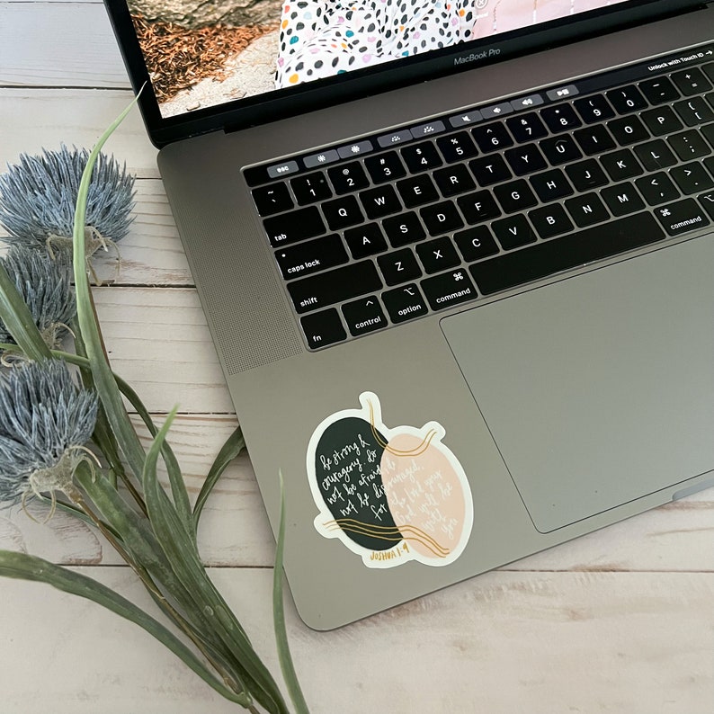 May include: A black and pink sticker with gold accents on a laptop. The sticker features a quote from Joshua 1:9, "Be strong and courageous. Do not be afraid; do not be discouraged, for the Lord your God will be with you wherever you go."
