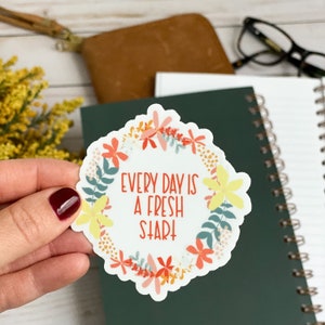 Peut inclure: Un autocollant blanc avec un motif de couronne de fleurs. La couronne est compos&eacute;e de fleurs jaunes, orange et roses. Le texte "Every day is a fresh start" est &eacute;crit en rouge &agrave; l'int&eacute;rieur de la couronne.