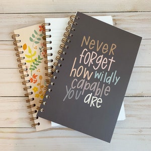 May include: Two spiral bound notebooks, one with a floral design and the other with the text "Never forget how wildly capable you are" in colourful lettering.