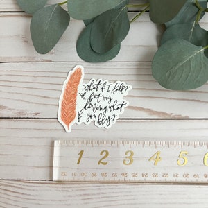 Op de afbeelding: Een sticker met een oranje veer en de tekst "what if I fall? oh but my darling, what if you fly?"
