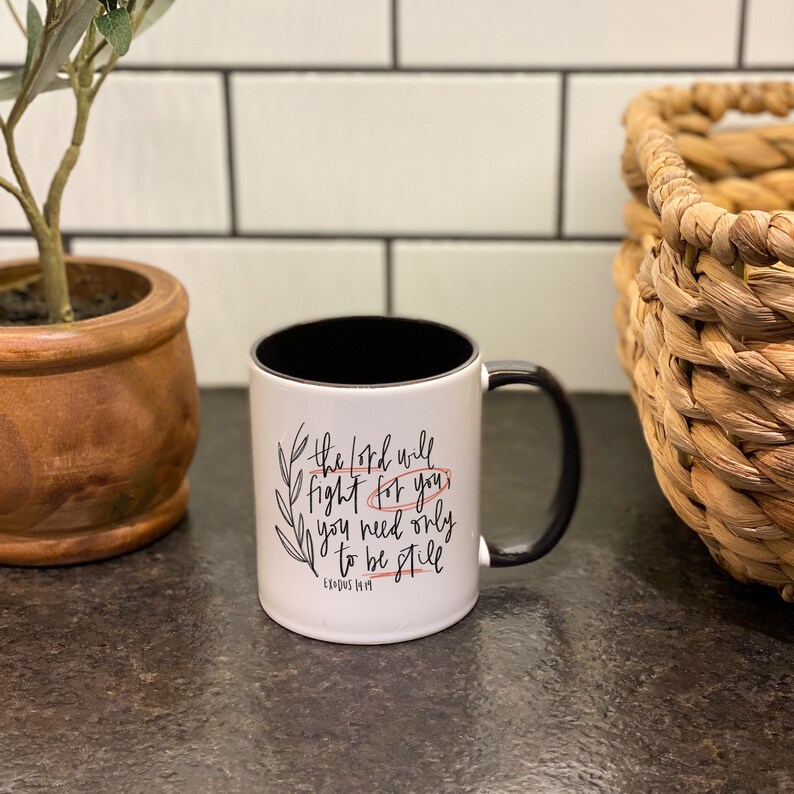 Op de afbeelding: Een witte keramische mok met een zwarte rand en handvat. De mok heeft een zwart-wit ontwerp met de tekst "The Lord will fight for you, you need only to be still. Exodus 14:14"