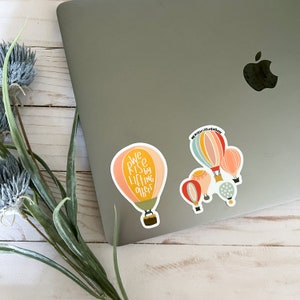 May include: A set of five hot air balloon stickers with a quote that reads "We rise by lifting others". The balloons are in shades of pink, yellow, blue, and red.