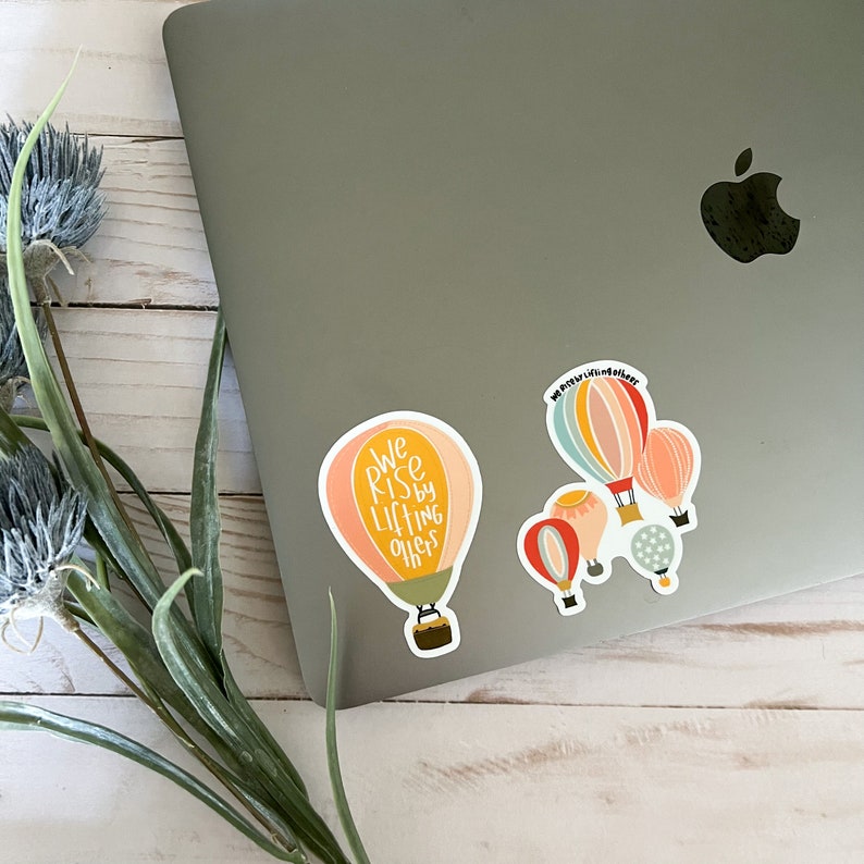 May include: A set of five hot air balloon stickers with a quote that reads "We rise by lifting others". The balloons are in shades of pink, yellow, blue, and red.