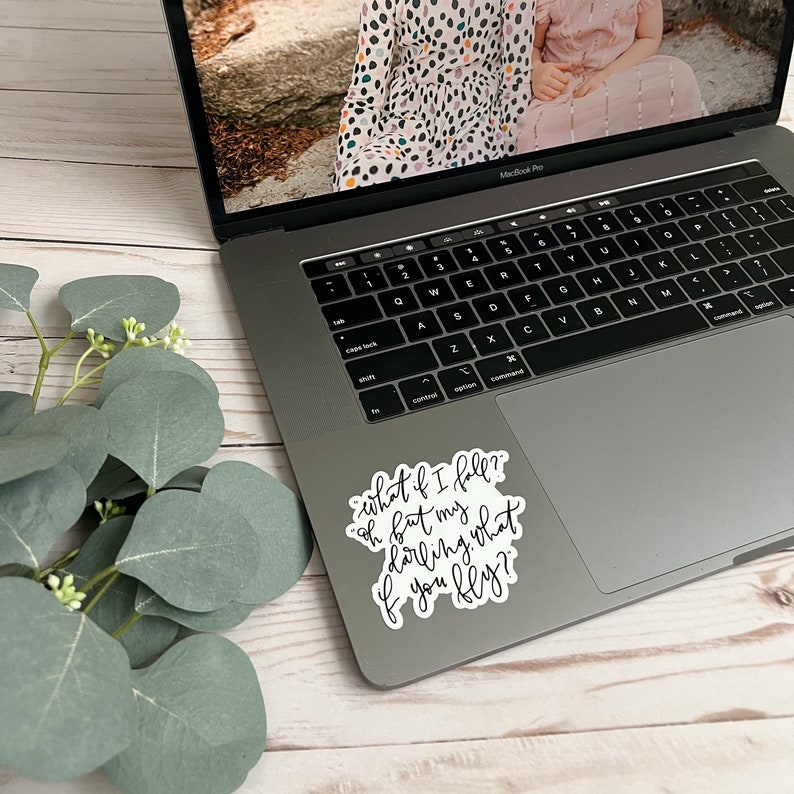 Op de afbeelding: Een witte laptop met een zwart toetsenbord en een sticker aan de rechterkant die zegt "what if I fall? oh but my darling, what if you fly?"