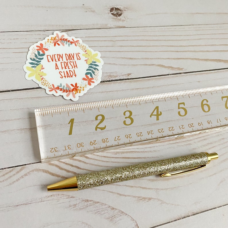 Peut inclure: Un stylo paillet&eacute; dor&eacute; avec une r&egrave;gle blanche en arri&egrave;re-plan. La r&egrave;gle comporte les chiffres de 1 &agrave; 7 imprim&eacute;s dessus. Un autocollant avec une couronne de fleurs et le texte "EVERY DAY IS A FRESH START" se trouve dans le coin sup&eacute;rieur gauche de l'image.