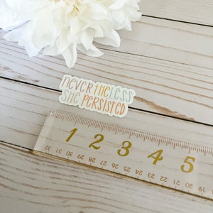 May include: A white sticker with the text "nevertheless she persisted" in colourful lettering. The sticker is on a wooden surface with a ruler next to it.
