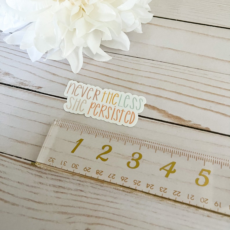 May include: A white sticker with the text "nevertheless she persisted" in colourful lettering. The sticker is on a wooden surface with a ruler next to it.
