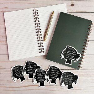 May include: Six black and white stickers with a silhouette of a woman's head and the text "Perhaps this is the moment for which you have been created". The stickers are on a light wood surface with two spiral notebooks and a pen.