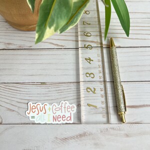 Puede incluir: Una pegatina con el texto "Jesus + Coffee = all I need" en letras de colores. La pegatina est&aacute; sobre una superficie de madera blanca con una regla y un bol&iacute;grafo dorado.