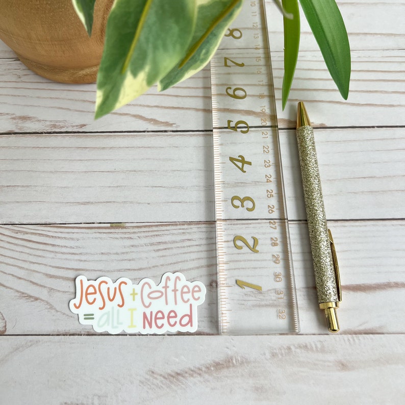 Puede incluir: Una pegatina con el texto "Jesus + Coffee = all I need" en letras de colores. La pegatina est&aacute; sobre una superficie de madera blanca con una regla y un bol&iacute;grafo dorado.