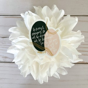 May include: A sticker with a black and pink background and gold accents. The sticker features a quote from the Bible, Joshua 1:9, which reads "Be strong and courageous. Do not be afraid; do not be discouraged, for the Lord your God will be with you."