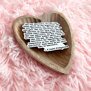 May include: A heart-shaped wooden dish with a white sticker featuring a quote by F. Scott Fitzgerald. The quote reads: "She was beautiful, but not like those girls in the magazines. She was beautiful, for the way she thought. She was beautiful, for the sparkle in her eyes when she talked about something she loved. She was beautiful, for her ability to make other people smile, even if she was sad. No, she wasn't beautiful for something as temporary as her looks. She was beautiful, deep down to her soul. She is beautiful."