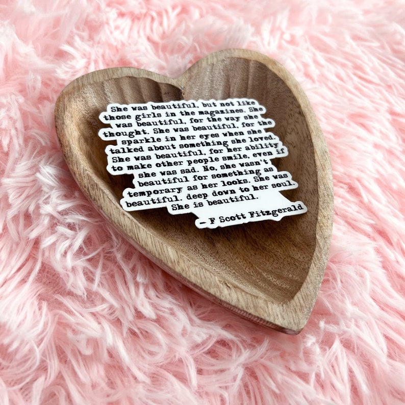 May include: A heart-shaped wooden dish with a white sticker featuring a quote by F. Scott Fitzgerald. The quote reads: "She was beautiful, but not like those girls in the magazines. She was beautiful, for the way she thought. She was beautiful, for the sparkle in her eyes when she talked about something she loved. She was beautiful, for her ability to make other people smile, even if she was sad. No, she wasn't beautiful for something as temporary as her looks. She was beautiful, deep down to her soul. She is beautiful."
