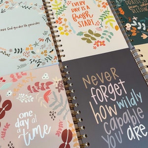 May include: A stack of five spiral bound journals with inspirational quotes and floral designs. The quotes include "Every day is a fresh start", "Never forget how wildly capable you are", and "One day at a time".
