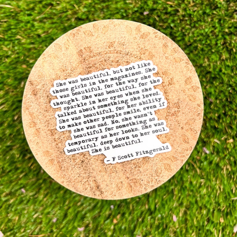 May include: A close-up of a round, cork-like coaster with a white sticker featuring a quote by F. Scott Fitzgerald. The quote reads: "She was beautiful, but not like those girls in the magazines. She was beautiful, for the way she thought. She was beautiful, for the sparkle in her eyes when she talked about something she loved. She was beautiful, for her ability to make other people smile, even if she was sad. No, she wasn't beautiful for something as temporary as her looks. She was beautiful, deep down to her soul. She is beautiful. - F. Scott Fitzgerald"