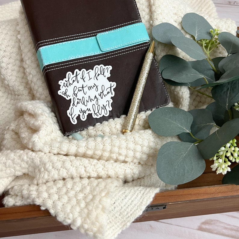 Op de afbeelding: Een witte sticker met zwarte kalligrafie met de tekst "what if I fall? oh but my darling, what if you fly?" De sticker is geplaatst op een bruin lederen dagboek met een turquoise lint.