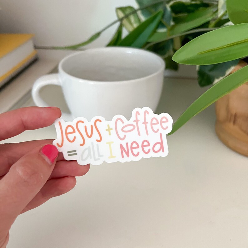 Puede incluir: Una pegatina blanca con el texto "Jesus + Coffee = all I need" en letras rosa, naranja y amarillo. La pegatina est&aacute; siendo sostenida por una mano.