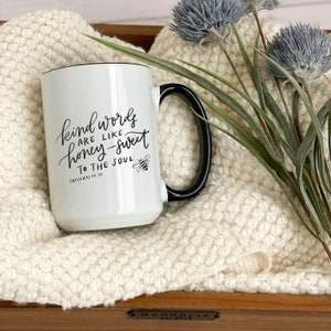 May include: White ceramic mug with a black handle. The mug has a black design that says "Kind words are like honey-sweet to the soul" with a bee illustration and the text "Proverbs 16:24".