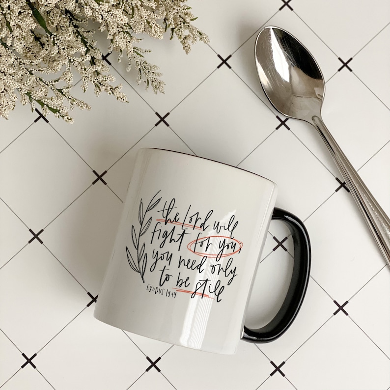 Op de afbeelding: Een witte keramische mok met een zwarte handgreep. De mok heeft een zwart-wit ontwerp met de tekst "The Lord will fight for you, you need only to be still" en de Bijbelverwijzing "Exodus 14:14".