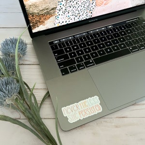 May include: A laptop computer with a sticker that reads "nevertheless she persisted" in colourful lettering. The sticker is on the right side of the trackpad.