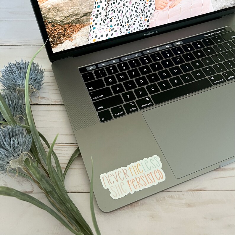 May include: A laptop computer with a sticker that reads "nevertheless she persisted" in colourful lettering. The sticker is on the right side of the trackpad.