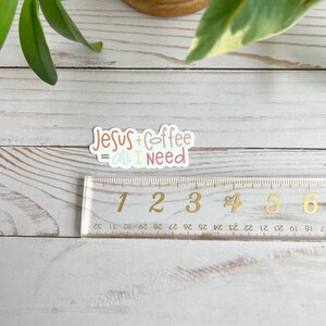 Puede incluir: Una pegatina con el texto "Jesus + Coffee = All I Need" en una fuente manuscrita colorida. La pegatina est&aacute; sobre una superficie de madera con una regla al lado.