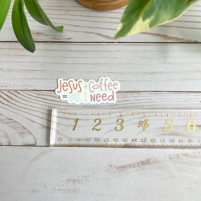 Puede incluir: Una pegatina con el texto "Jesus + Coffee = All I Need" en una fuente manuscrita colorida. La pegatina est&aacute; sobre una superficie de madera con una regla al lado.