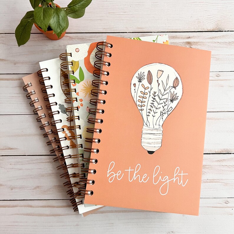 Puede incluir: Una pila de cuatro cuadernos de espiral con una cubierta de color melocot&oacute;n. La portada del cuaderno superior presenta una bombilla con dise&ntilde;os florales en su interior y el texto "be the light".