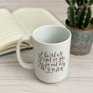 Op de afbeelding: Witte keramische mok met zwarte kalligrafie die "De Heer zal voor je vechten, je hoeft alleen maar stil te zijn" luidt, met de Bijbelverwijzing "Exodus 14:14" in kleiner lettertype eronder. De mok staat op een licht houten oppervlak met een boek en een cactus in een pot op de achtergrond.