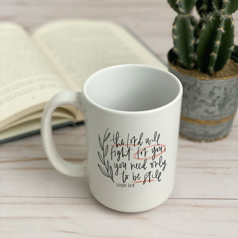 Op de afbeelding: Witte keramische mok met zwarte kalligrafie die "De Heer zal voor je vechten, je hoeft alleen maar stil te zijn" luidt, met de Bijbelverwijzing "Exodus 14:14" in kleiner lettertype eronder. De mok staat op een licht houten oppervlak met een boek en een cactus in een pot op de achtergrond.