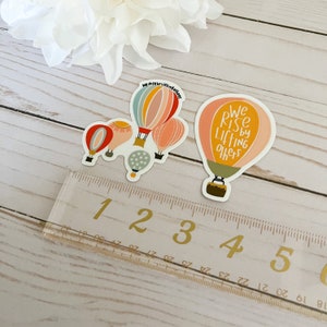May include: Two hot air balloon stickers. One sticker features a cluster of four hot air balloons with the text "We rise by lifting others". The other sticker features a single hot air balloon with the text "We rise by lifting others".