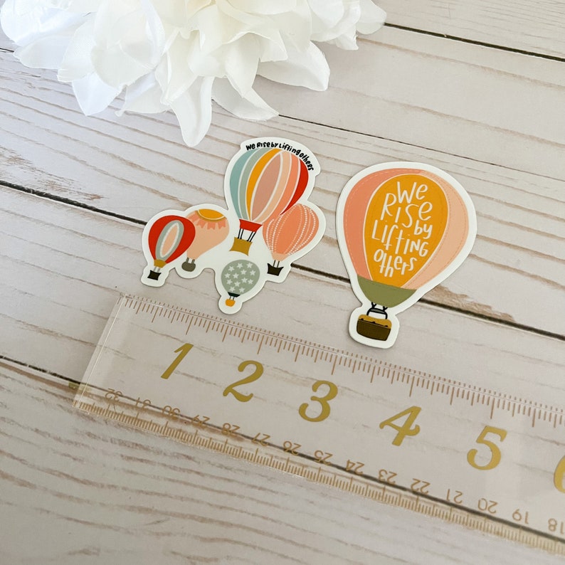 May include: Two hot air balloon stickers. One sticker features a cluster of four hot air balloons with the text "We rise by lifting others". The other sticker features a single hot air balloon with the text "We rise by lifting others".