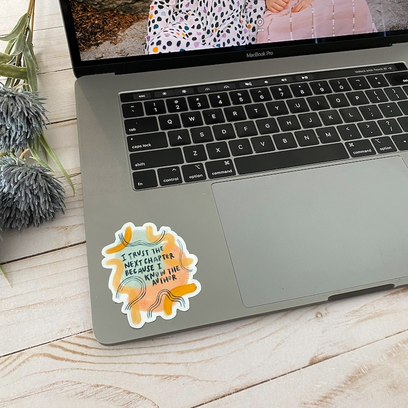 May include: A sticker with a watercolor background and yellow, orange, and blue accents. The sticker features the text "I trust the next chapter because I know the author."
