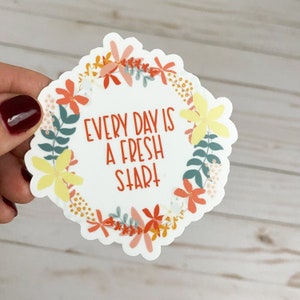 Peut inclure: Un autocollant blanc avec un motif de couronne de fleurs. La couronne est compos&eacute;e de fleurs orange, jaunes et roses. Le texte "EVERY DAY IS A FRESH START" est &eacute;crit en rouge &agrave; l'int&eacute;rieur de la couronne.