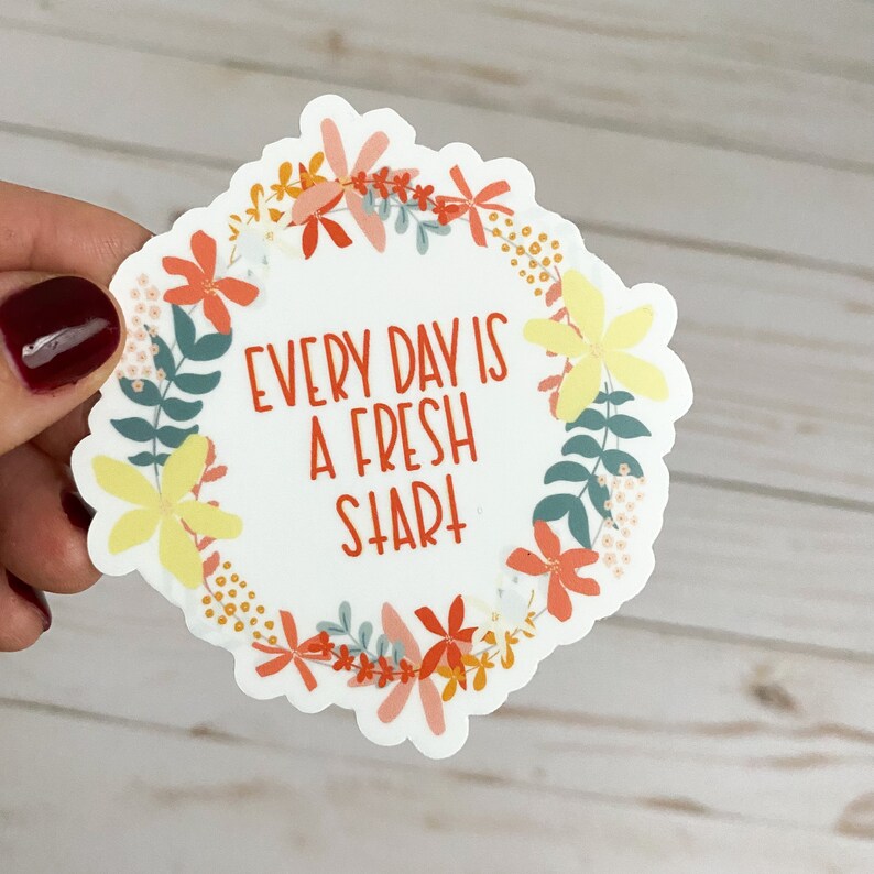 Peut inclure: Un autocollant blanc avec un motif de couronne de fleurs. La couronne est compos&eacute;e de fleurs orange, jaunes et roses. Le texte "EVERY DAY IS A FRESH START" est &eacute;crit en rouge &agrave; l'int&eacute;rieur de la couronne.