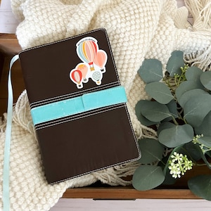 May include: A brown leather journal with a turquoise ribbon closure. A sticker with colourful hot air balloons and the text "We're Just Like Hot Air Balloons" is on the front cover.