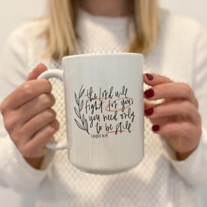 Op de afbeelding: Een witte keramische mok met een zwart en oranje ontwerp. De mok heeft de tekst "The Lord will fight for you, you need only to be still" en "Exodus 14:14".
