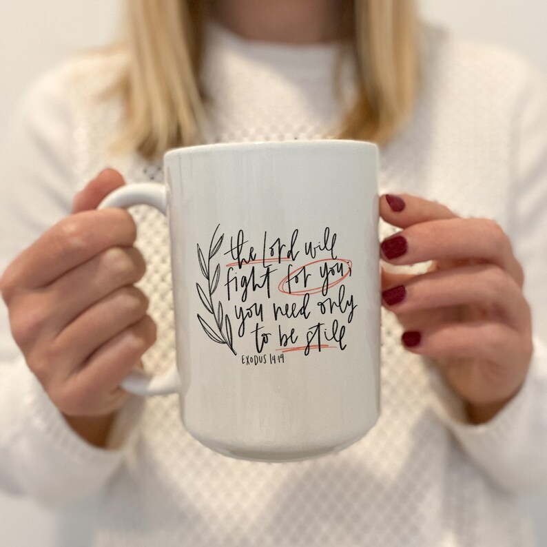 Op de afbeelding: Een witte keramische mok met een zwart en oranje ontwerp. De mok heeft de tekst "The Lord will fight for you, you need only to be still" en "Exodus 14:14".