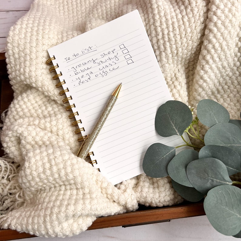 Puede incluir: Un cuaderno blanco con una encuadernaci&oacute;n en espiral dorada y una lista de tareas escritas en la primera p&aacute;gina. La lista incluye ir de compras, estudiar la Biblia, clase de yoga y la oficina de correos. Un bol&iacute;grafo de purpurina dorado est&aacute; sobre el cuaderno.