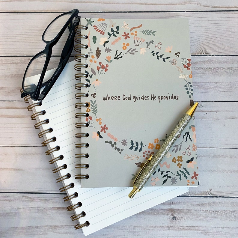 May include: A gray journal with a floral design and the text "where God guides He provides". The journal has a spiral binding and is open to a blank page. A gold pen with glitter is lying on top of the journal.